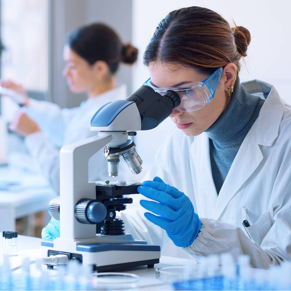 Woman in lab coat and goggles looking through microscope